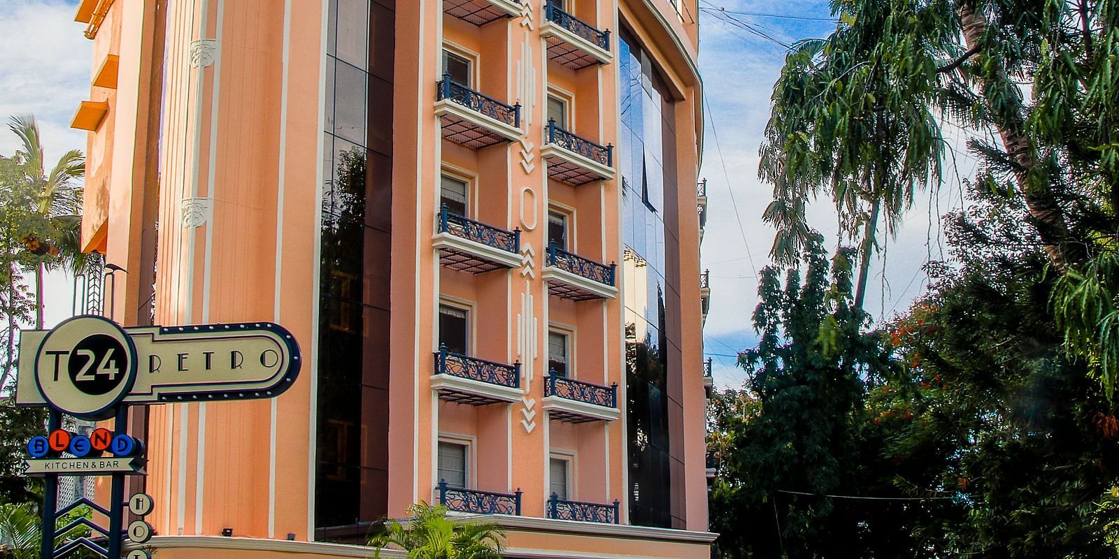 The exterior of the T24 Retro, a 4-star boutique hotel in Mumbai, India. The peach-colored building has a retro 70s style, with black balconies and large windows. Palm trees and other greenery line the street in front. 